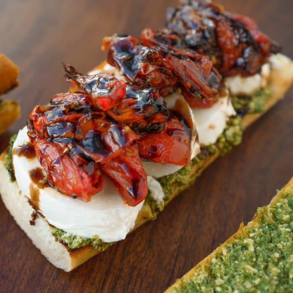 Three bruschetta pieces with pesto, mozzarella, roasted tomatoes, and balsamic glaze on a wooden board.