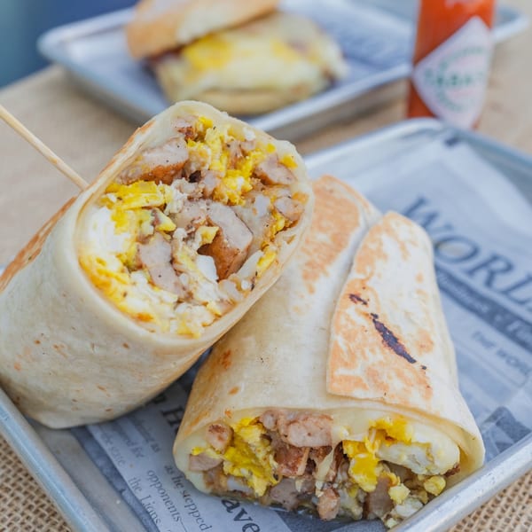 Two breakfast burritos filled with scrambled eggs and meat, served on a metal tray with newspaper.