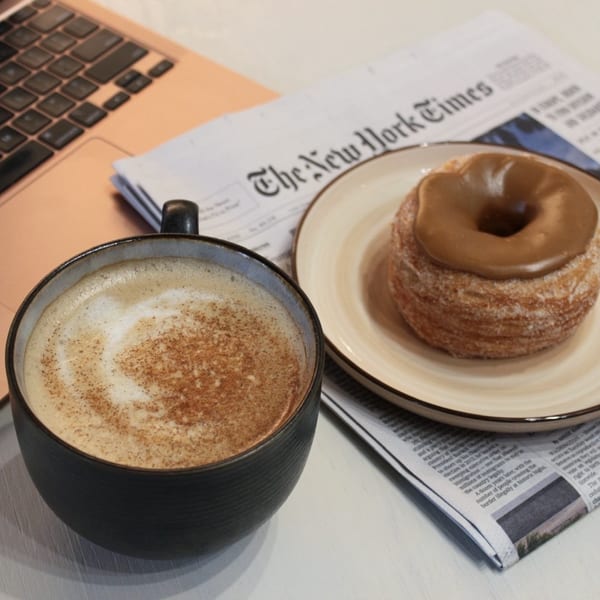 A latte with cinnamon, a cronut with caramel glaze, and a newspaper on a table.