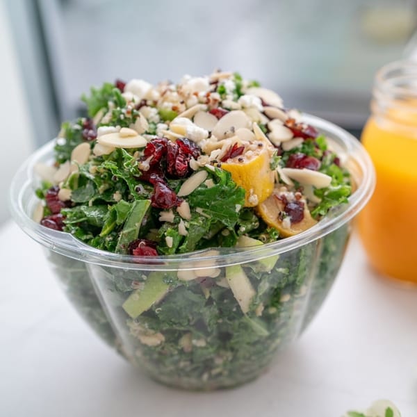 A vibrant kale salad with cranberries, almonds, and feta cheese in a clear plastic bowl.
