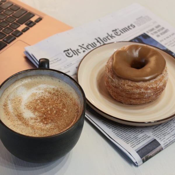 A coffee with latte art and a cronut on a plate next to a newspaper and laptop.